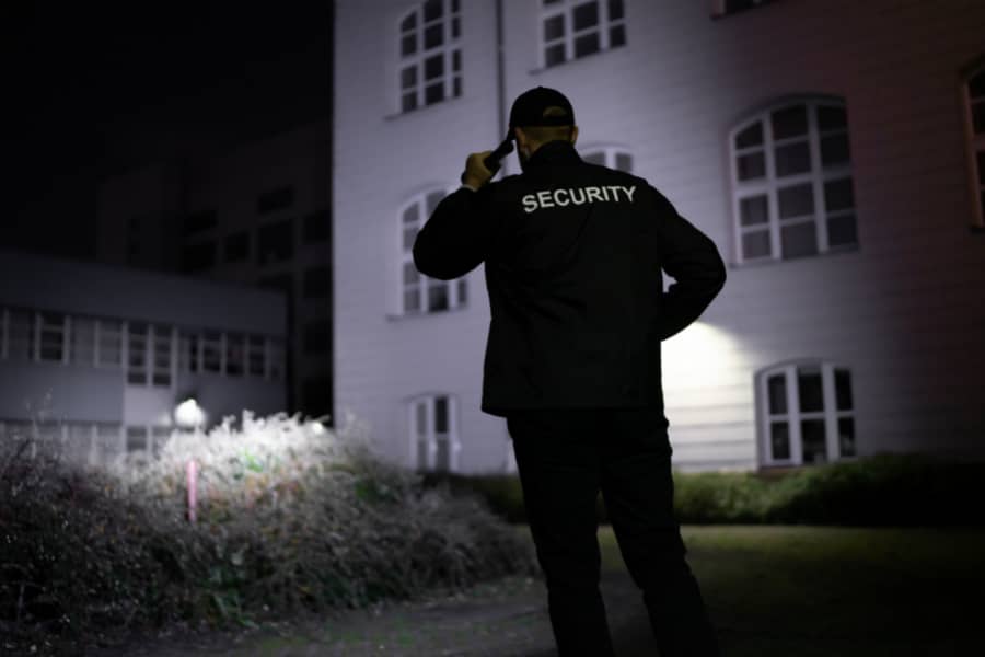 Manned Security guard on night patrols arounds grounds of business shining torch at shrubbery and grass area